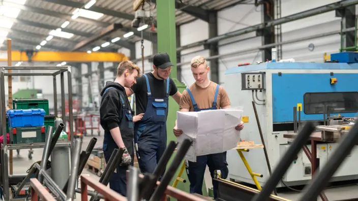 Drei Handwerker stehen in einer Fabrik und schauen gemeinsam auf einen Bauplan. 