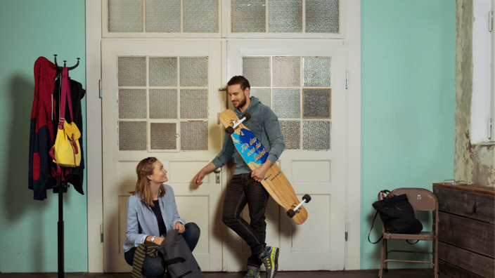 junges Paar lehnt an der Zimmertür einer Altbauwohnung, sie packt ihren Rucksack, er hält lässig ein Longboard im Arm