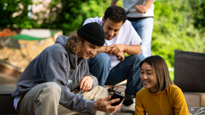 drei junge Leute sitzen in einem Skatepark und blicken auf ein Smartphone