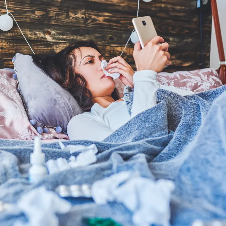 Frau liegt krank mit Schnupfen im Bett und betätigt ihr Smartphone