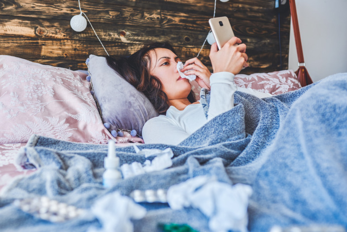 Frau liegt krank mit Schnupfen im Bett und betätigt ihr Smartphone