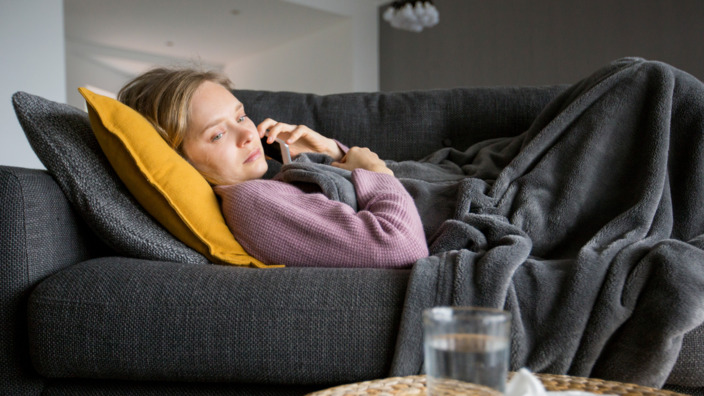 Frau liegt krank auf Sofa mit Telefon in der Hand