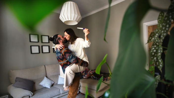 Mann hebt seine Frau hoch. Frau hält Schwangerschaftstest in der Hand.