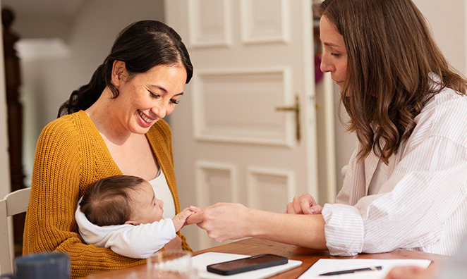 Mutter mit Baby sitzen am Tisch, zweite Person sitzt gegenüber, beide lächeln.