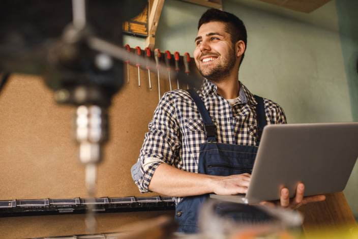 Junger Mann sitzt mit Laptop in der Hand an einer Werkbank