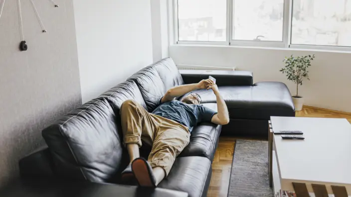 Junger Mann schaut in seine Smartphone auf einer Couch liegend