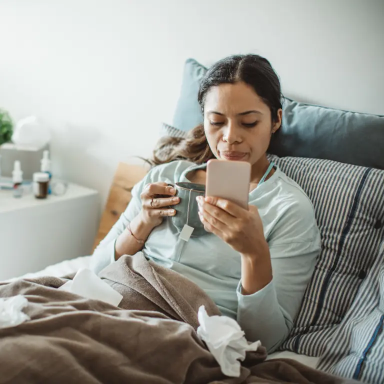 Eine Frau liegt krank im Bett und hält in ihrer Hand ein Smartphone.