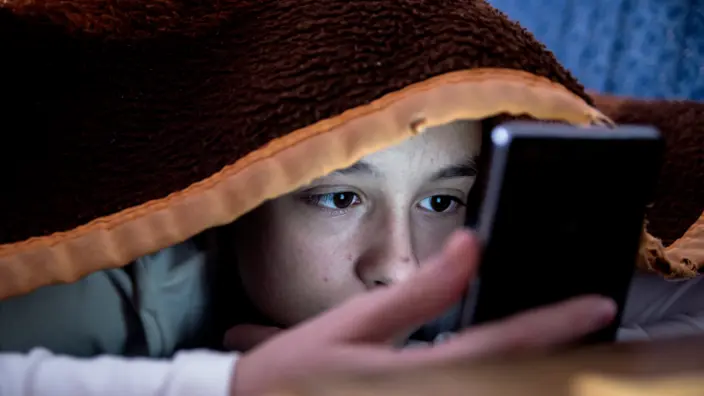 Kind schaut, versteckt unter eine Decke, auf das Display eines Smartphones.