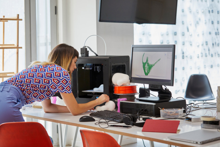 Eine Frau beugt sich über einen Schreibtisch mit einem 3D-Drucker, auf einem Bildschirm ist ein digitales Modell zu sehen.