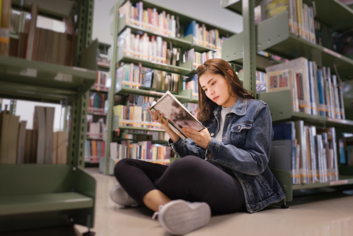 Junge Frau sitzt mit aufgeschlagenen Buch in der Hand auf dem Boden vor vollen Bücherregalen