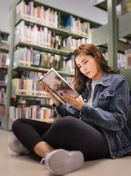 Junge Frau sitzt mit aufgeschlagenen Buch in der Hand auf dem Boden vor vollen Bücherregalen