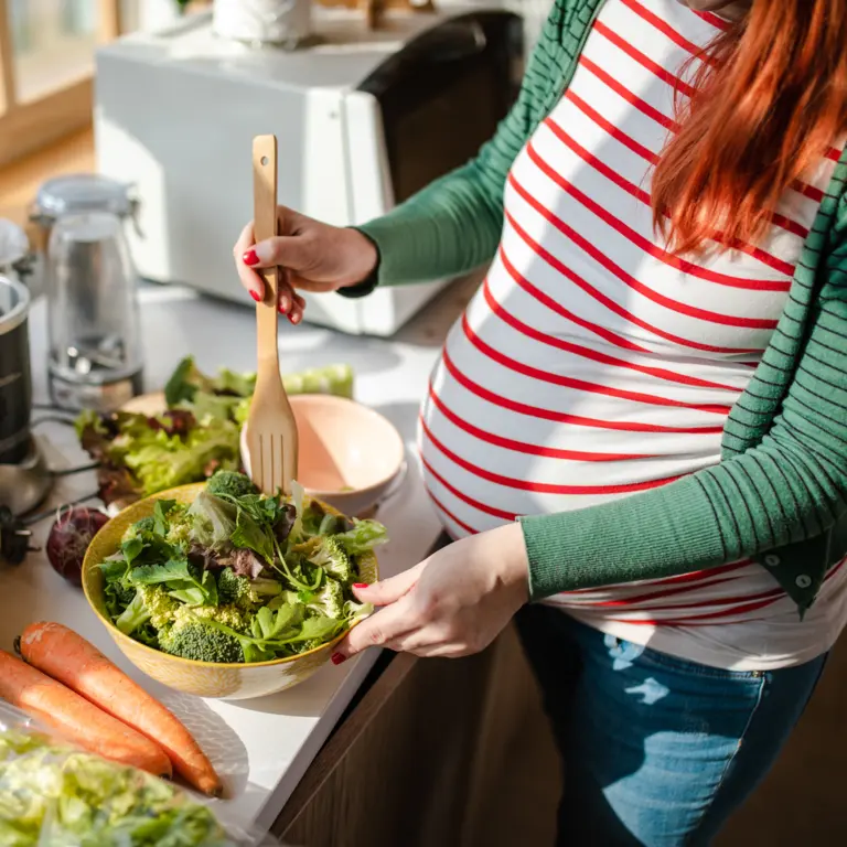 Schwangere bereitet Salat zu auf einer Küchenzeile zu, auf der viel Obst und Gemüse ausliegt