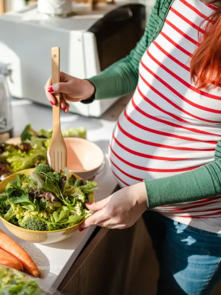 Schwangere bereitet Salat zu auf einer Küchenzeile zu, auf der viel Obst und Gemüse ausliegt