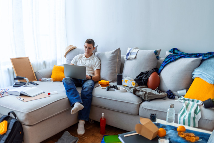 Teenager sitzt auf dem Sofa umgeben von Durcheinander und Dingen.