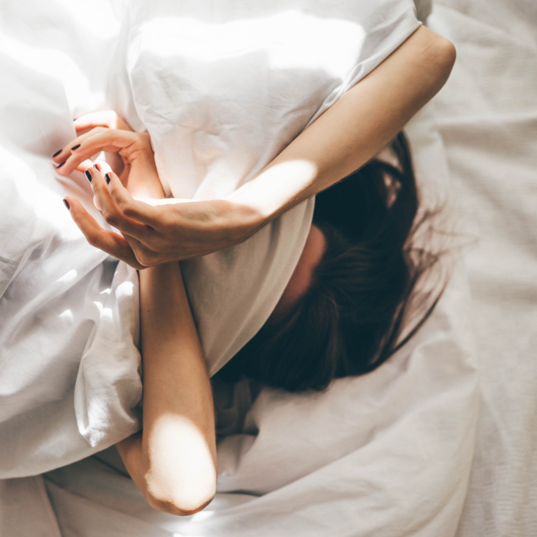 Eine Frau liegt entspannt im Bett und hat siech die Decke über das Gesicht gezogen