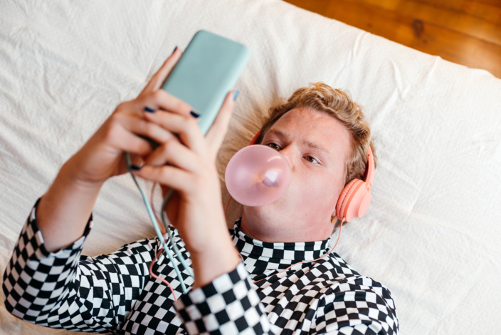 Junger Mensch, der mit Smartphone in der Hand auf dem Bett liegt und eine Kaugummiblase macht.