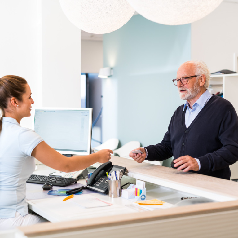 Alter Mann steht am Empfang in Arztpraxis und spricht mit einer Arzthelferin