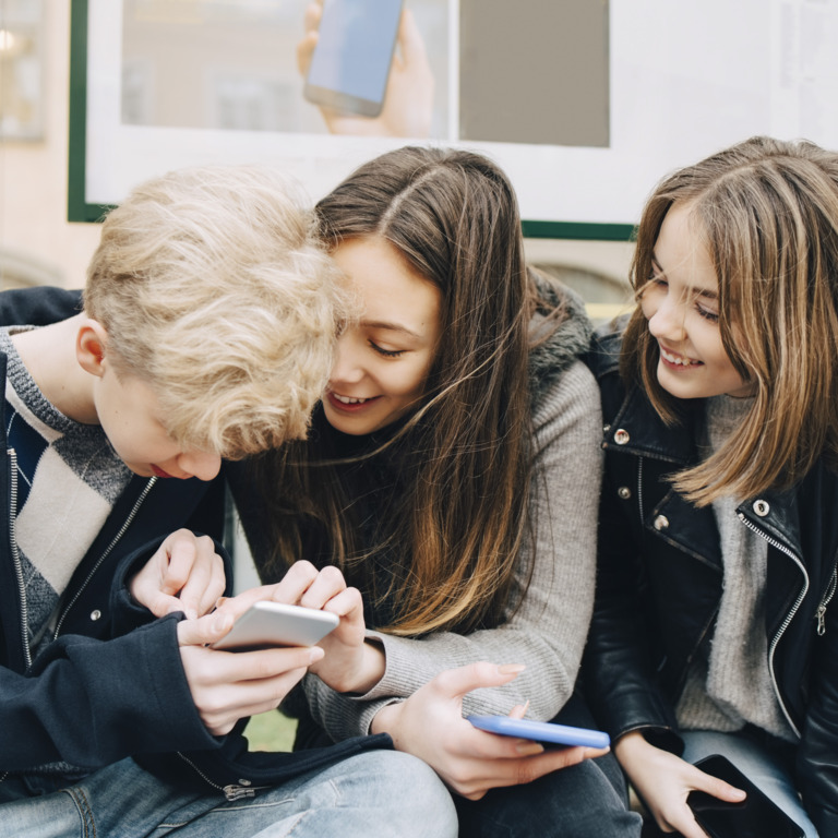 Drei Teenager sitzen an einer Bushaltestelle und schauen gemeinsam auf ihre Smartphones