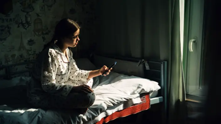 Ein Mädchen sitzt im dunklen Schlafzimmer auf dem Bett und hält ein Handy in der Hand