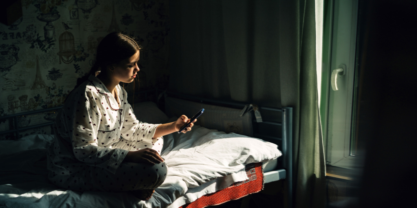 Weiblicher Teenager sitzt auf seinem Bett und schaut nachdenklich auf sein Smartphone.