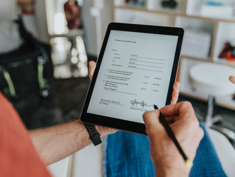 Auf einem Tablet wird ein medizinisches Formular unterschrieben. Man sieht nur die Hände der Person, die sich in einem Behandlungszimmer befindet.