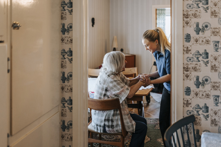 Pflegekraft gibt alter Dame die Hände. Die Dame sitzt auf einem Stuhl in ihrem Wohnzimmer, die Pflegekraft steht vor ihr.