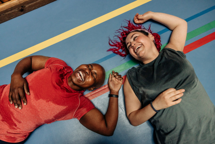 Zwei Frauen liegen lachend und schwitzend auf einem Sporthallenboden