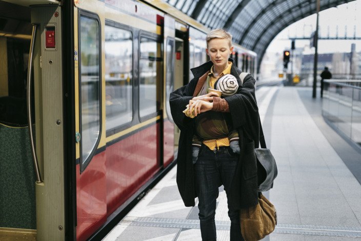 Eine Frau steht mit einem Baby auf dem Arm am Bahnsteig neben einem Zug und schaut auf die Uhr.