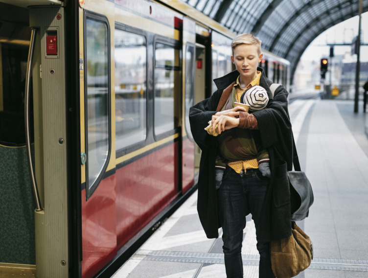 Eine Frau steht mit einem Baby auf dem Arm am Bahnsteig neben einem Zug und schaut auf ihre Armbanduhr.