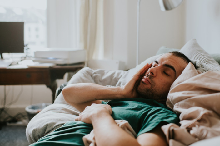 Mann liegt noch verschlafen im Bett und streicht sich über das Gesicht