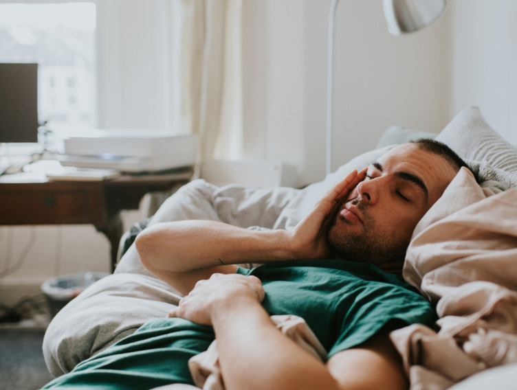 Mann liegt noch verschlafen im Bett und streicht sich über das Gesicht