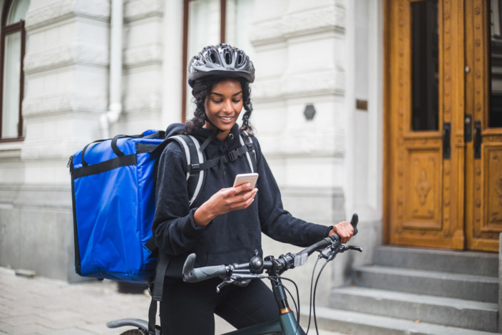 Frau vom Lieferdienst schaut auf ihr Handy, während sie mit ihrem Fahrrad vor einer Haustür steht