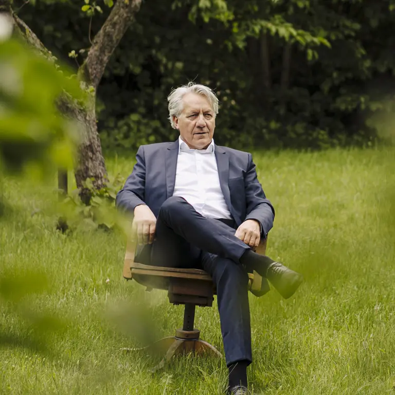 Reifer Mann im Anzug sitzt auf einem Schreibtischstuhl im Garten – Symbol für Nachfolgesuche
