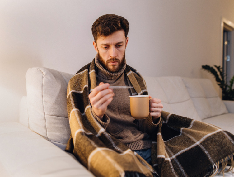 Mann sitzt mit Tasse und Fieberthermometer in der Hand auf dem Sofa. 