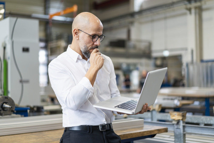 Mann steht in der Werkstatt und schaut auf seinen Laptop, den er in der linken Hand hält.