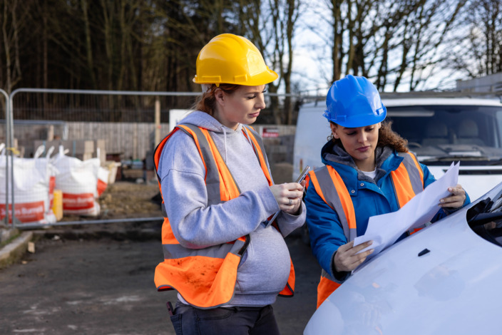 Schwangere mit Schutzhelm steht auf einer Baustelle und unterhält sich mit einer anderen Frau mit Helm