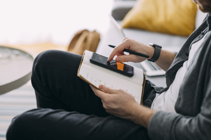 Mann sitzt mit Buch und Handy auf dem Schoß auf einem Sofa.