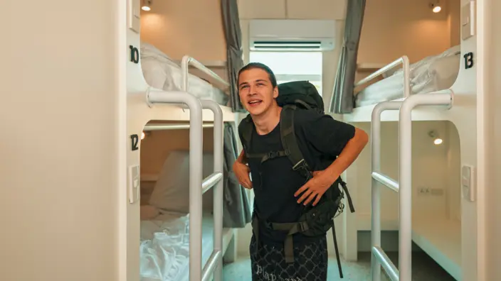 Junger Mann steht mit seinem Rucksack in einem Zimmer mit Stockbetten