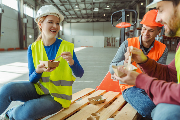 Arbeitnehmerin mit Helm und Weste isst Mittagessen mit Kollegen