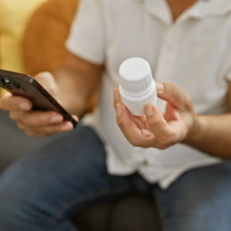 Ein Mann hält in einer Hand sein Smartphone und in der anderen eine Medikamentendose. 