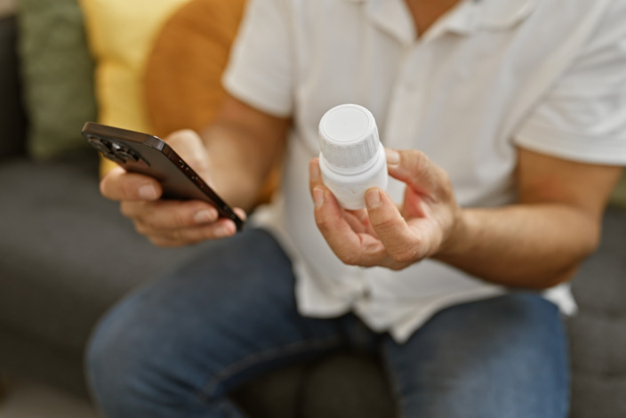 Ein Mann hält in einer Hand sein Smartphone und in der anderen eine Medikamentendose. 