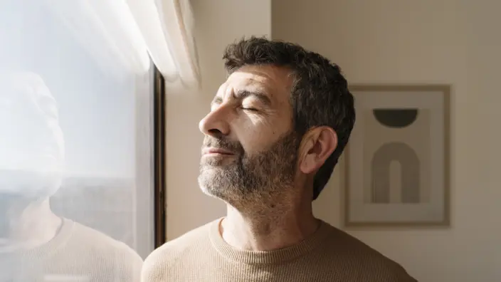 Mann steht mit geschlossenen Augen vor einem Fenster