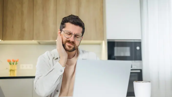 Ein Mann sitzt vor seinem Laptop und hält sich seinen schmerzenden Kiefer.