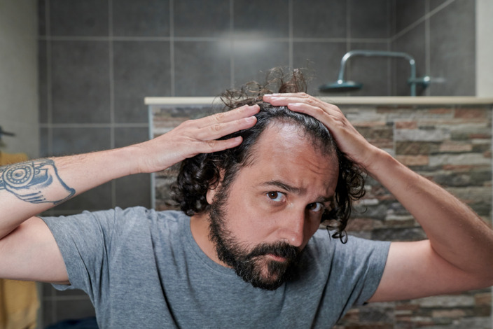 Tättowierter Mann steht in einem Bad und sieht sich seine Haare an. 