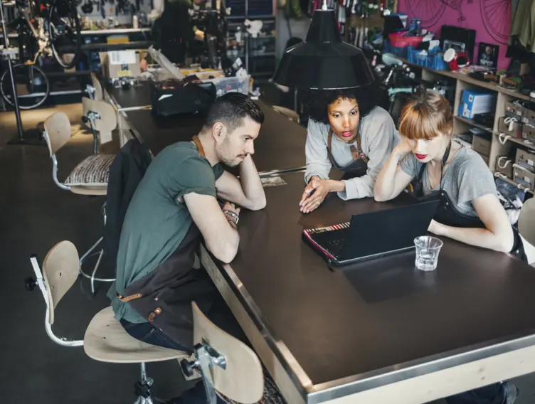 Eine Gruppe von zwei Handwerkerinnen und einem Handwerker sitzen an einem Tisch und schauen in einen Laptop. 