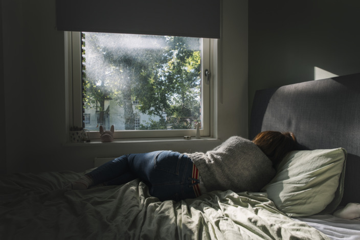 Frau liegt mit Rücken zur Kamera im Bett und schaut aus dem Fenster, wo die Sonne sich in Bäumen bricht.
