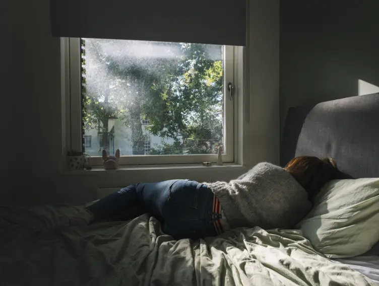 Frau liegt mit Rücken zur Kamera im Bett und schaut aus dem Fenster, wo die Sonne sich in Bäumen bricht.