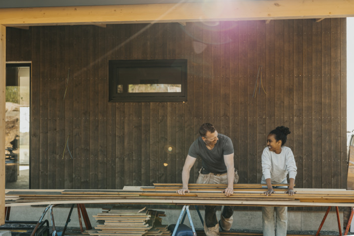 Vater und Tochter arbeiten gemeinsam an Holzlatten vor einer Holzfassade in der Sonne
