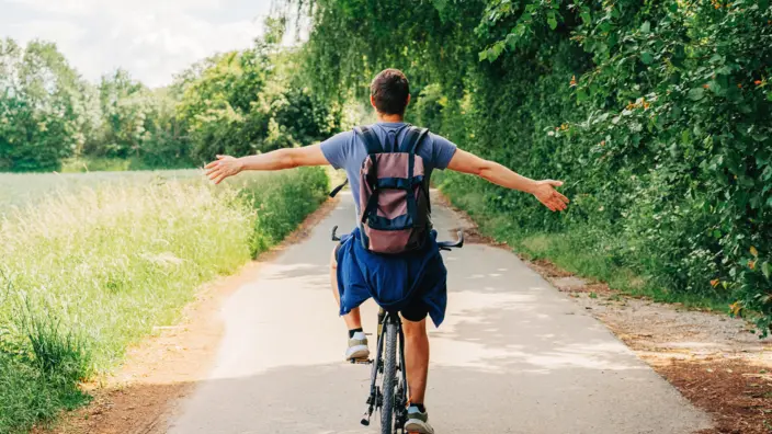 Mann fährt auf einem Fahrrad in einer Sommerlandschaft und streckt die Arme seitlich aus.