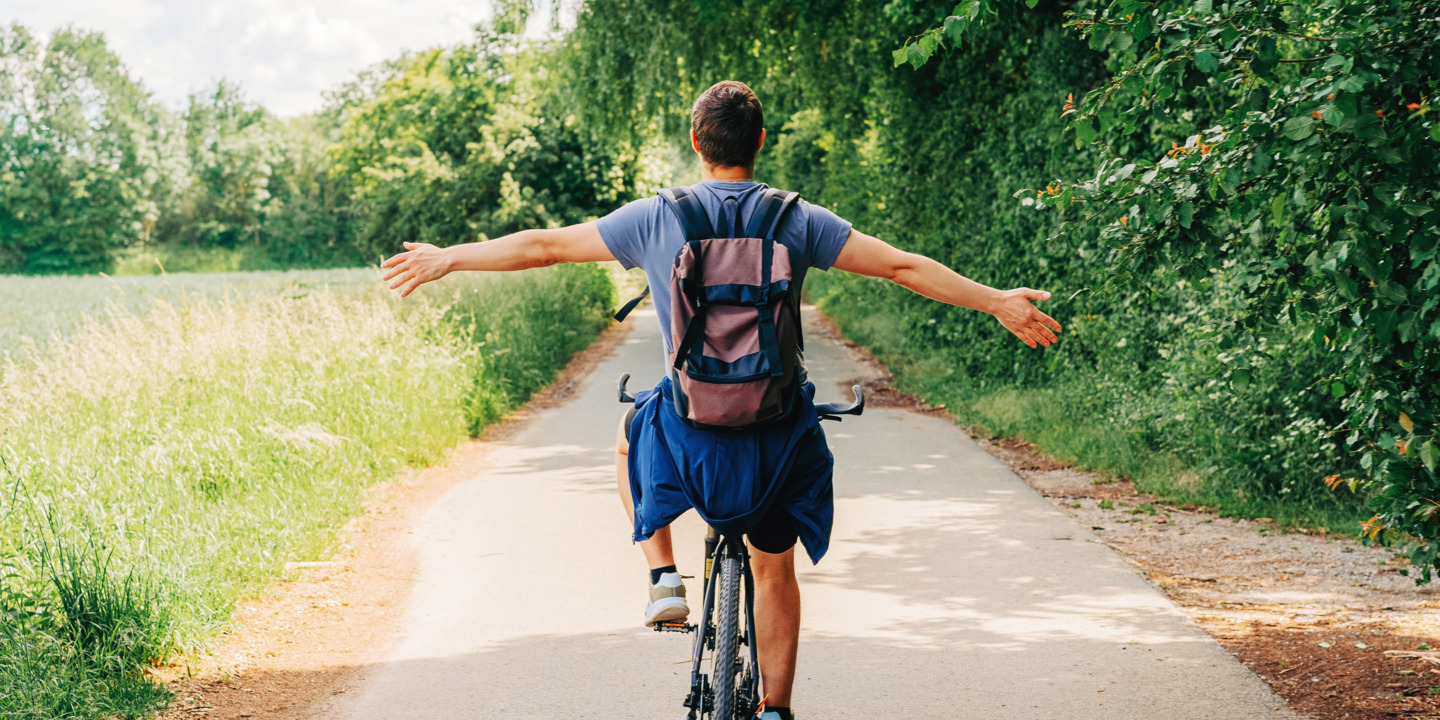 Mann fährt auf einem Fahrrad in einer Sommerlandschaft und streckt die Arme seitlich aus.